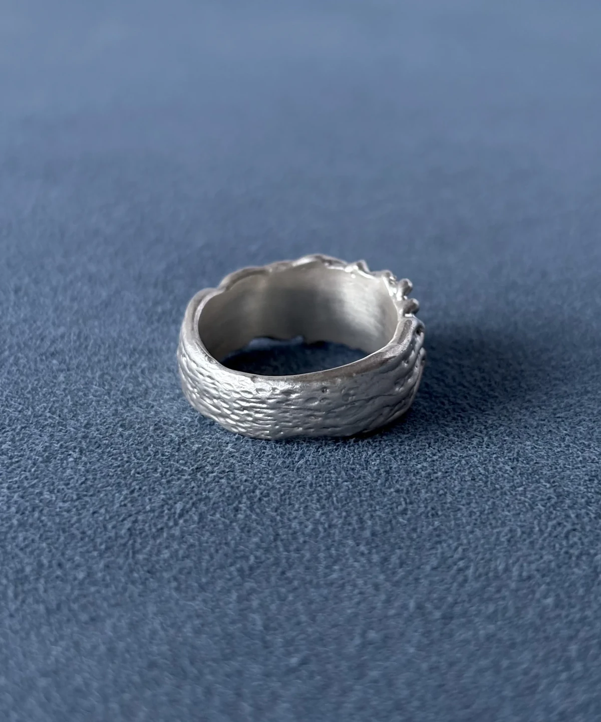 Back view of a wide silver ring with flowing surface texture and the inner band visible on blue fabric.