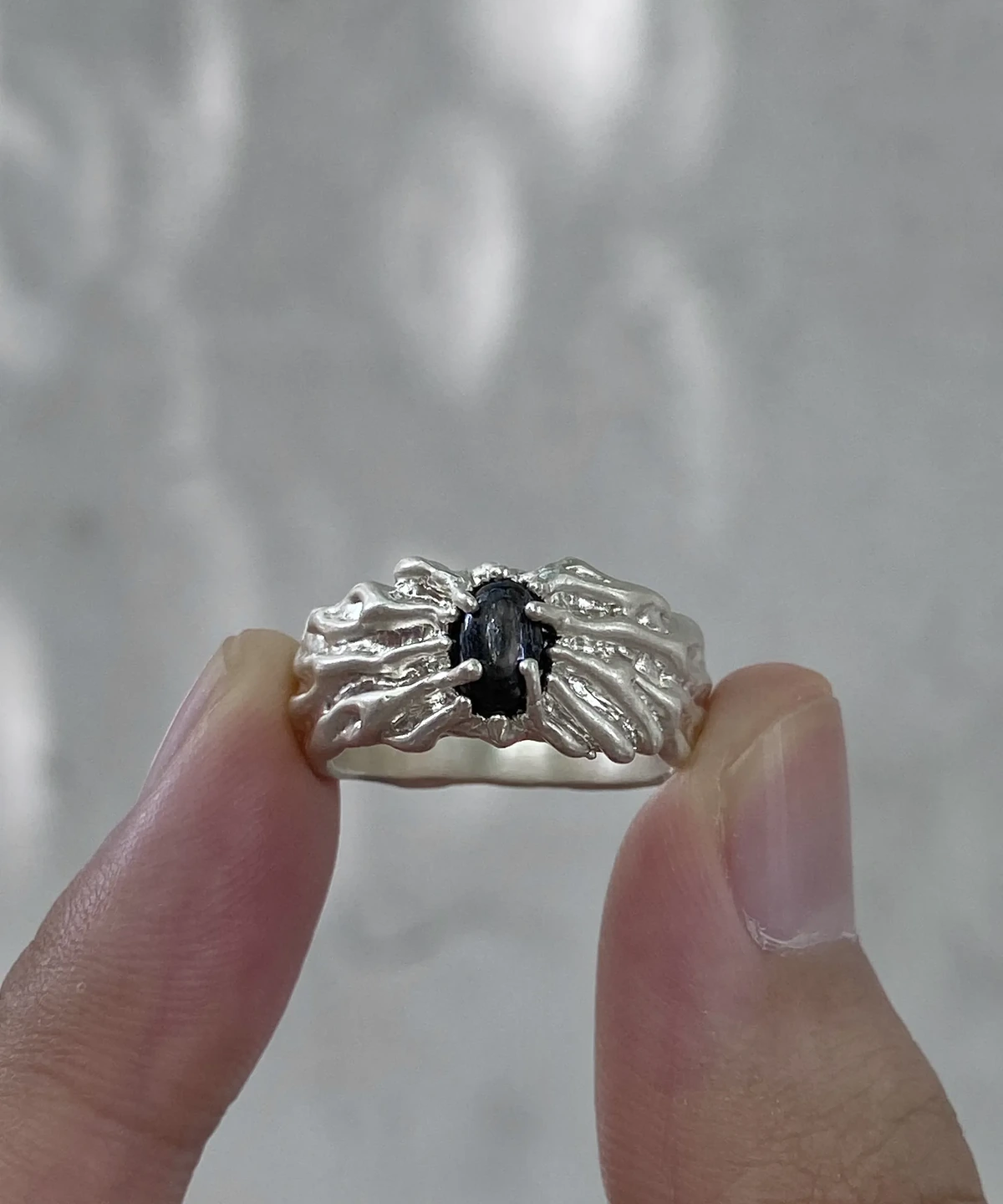 Hypersthene ring with an oval black stone and flowing silver ridges, held between two fingers against a grey background.