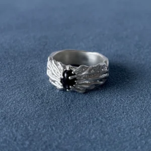 Wide silver ring with a dark oval centre stone and flowing raised texture, resting on blue fabric.