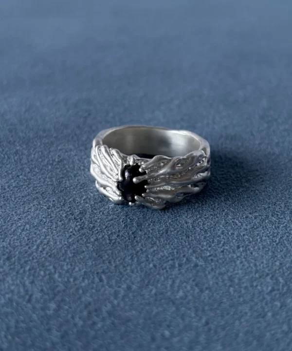 Wide silver ring with a dark oval centre stone and flowing raised texture, resting on blue fabric.