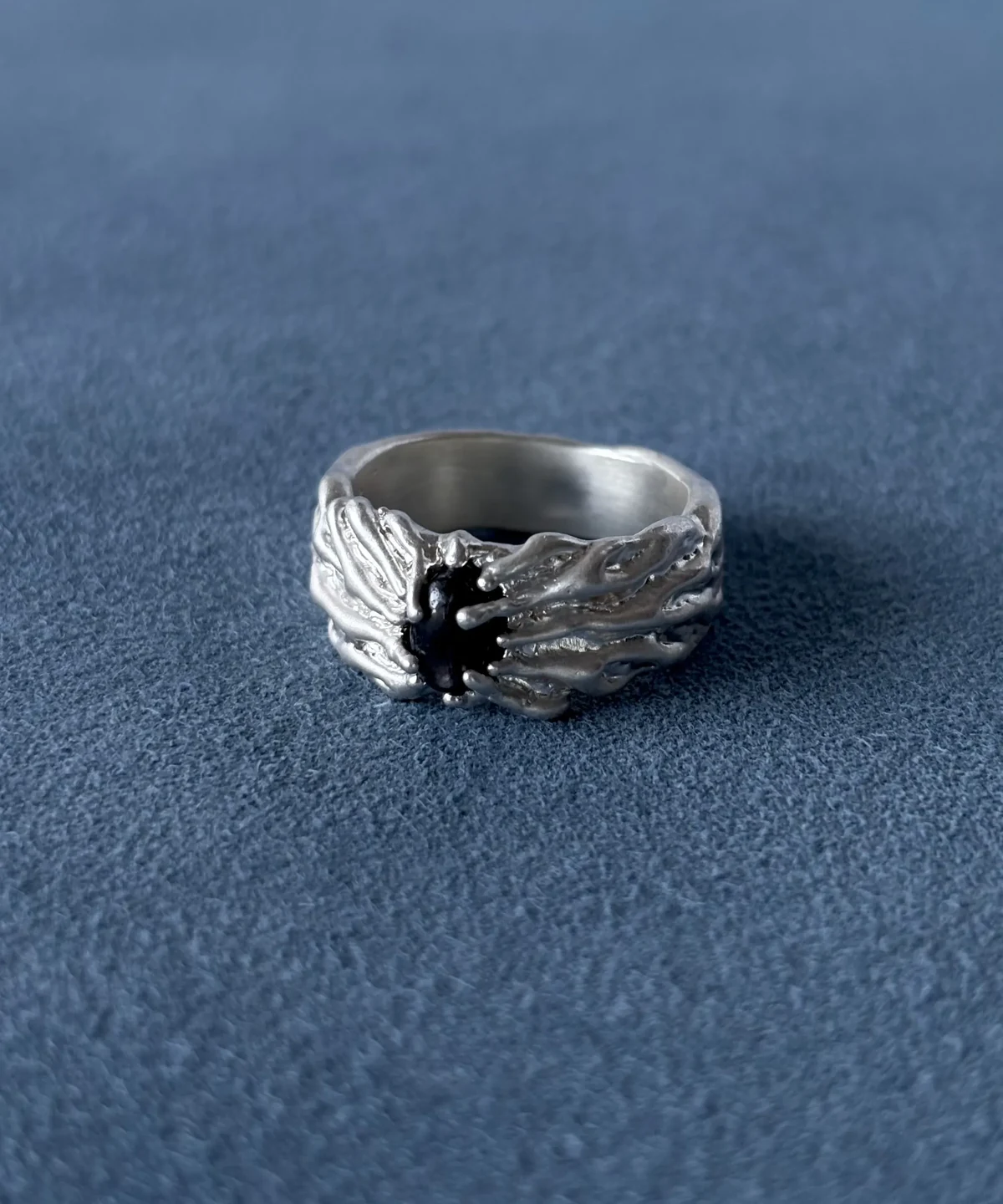 Wide silver ring with a dark oval centre stone and flowing raised texture, resting on blue fabric.