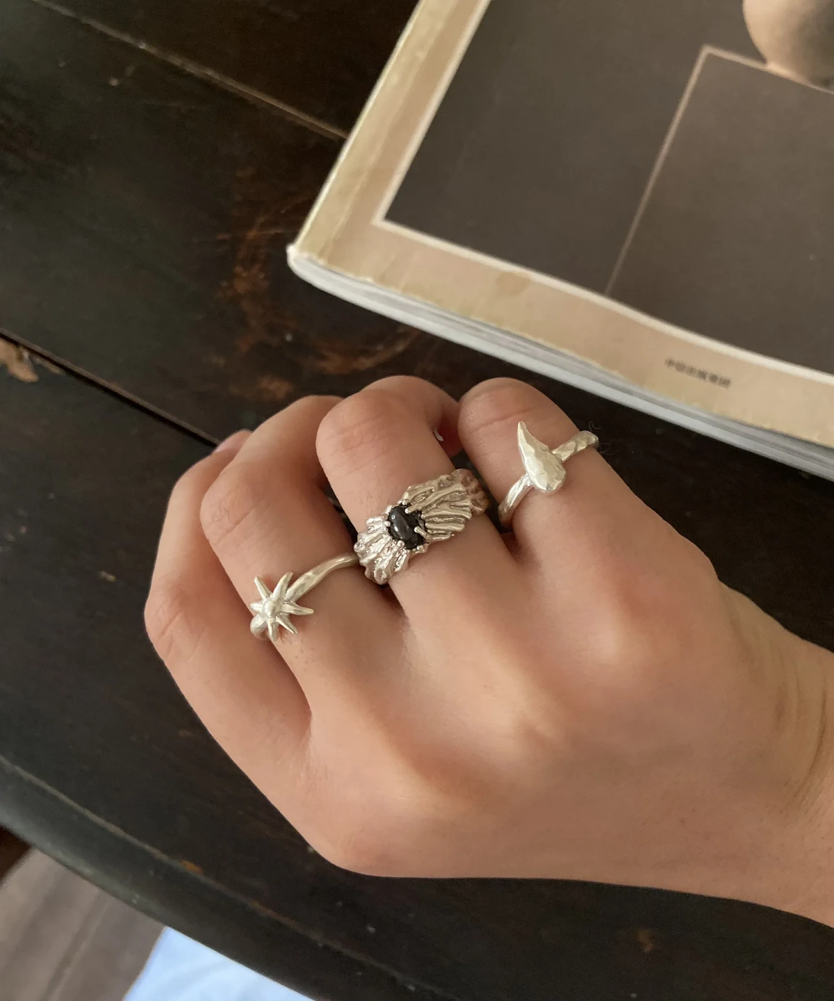 Black gemstone silver ring worn between a star ring and a fishtail ring beside an open book.