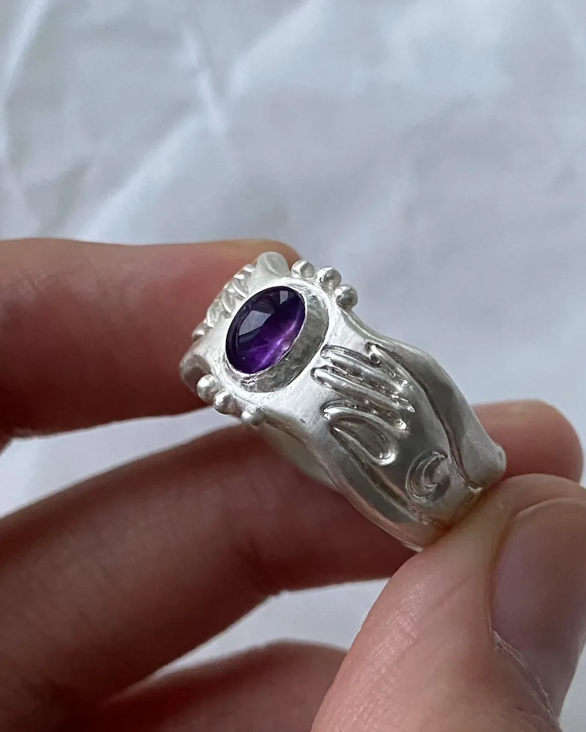 A Pocket of Cosmos 999 Silver Amethyst Ring A Pocket of Cosmos 999 silver ring held between fingers, with violet amethyst and sculpted hand and crescent details