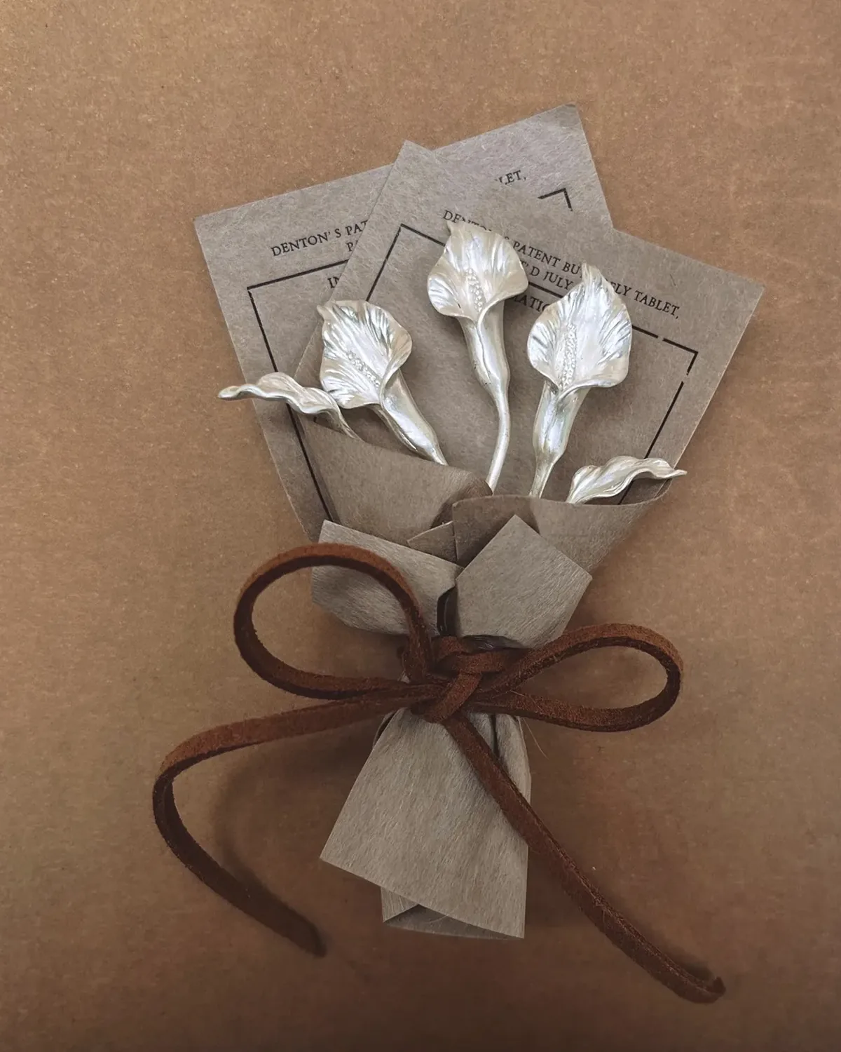 Miniature Silver Calla Lily Bouquet Three sculpted silver calla lilies tied into a miniature bouquet on a warm paper background
