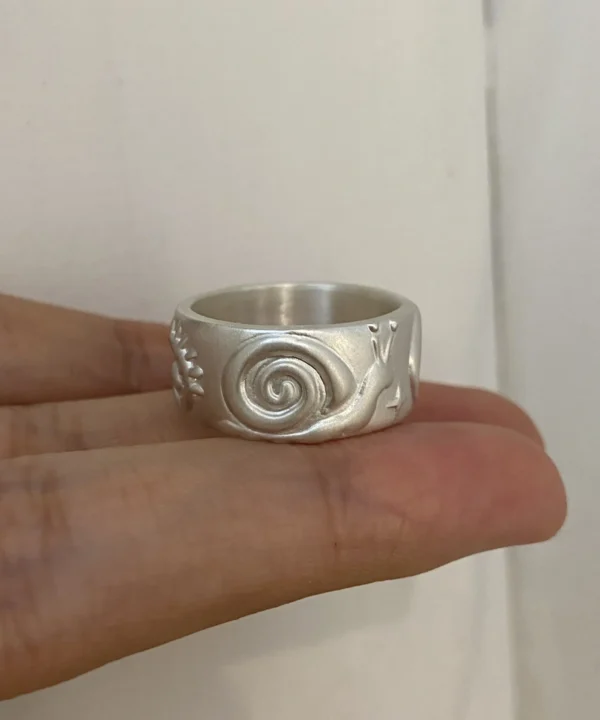 Silver snail ring with a matte wide band, raised spiral shell and antennae, held across the fingers.