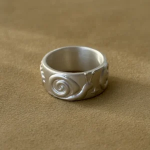 Silver snail ring with a raised spiral shell and matte wide band on ochre fabric.
