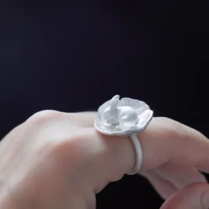 Side view of a rabbit resting in a cupped flower on a silver ring, worn against a dark background.